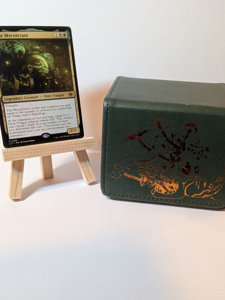 The MTG card "The Mycrotyrant" on a small wooden easel next to a green and foil gold deck bock with mushroom imagery. 