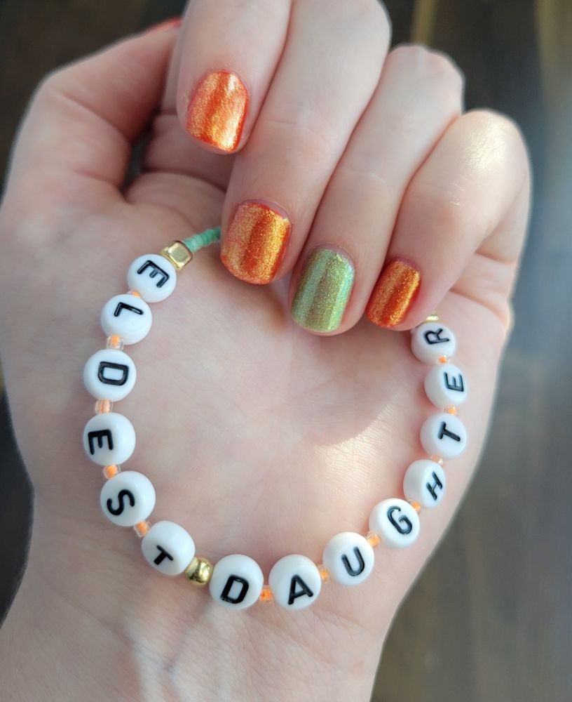 Glittery nails in orange with a mint accent finger (ring finger) grasping an orange and mint with gold accent bracelet reading ELDEST DAUGHTER
