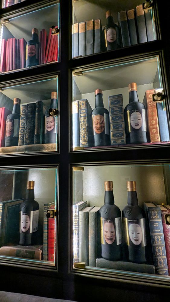 Shelves with books and wine bottles on which different pictures of Japanese Noh masks are featured
