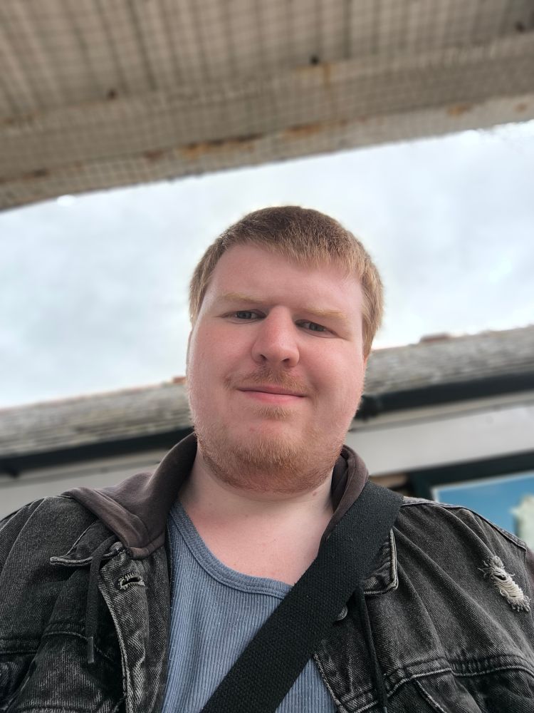 Selfie taken outdoors of a white, masc presenting person with short ginger hair wearing a denim hoodie with a blue shirt under and a black bag strap crossing diagonally