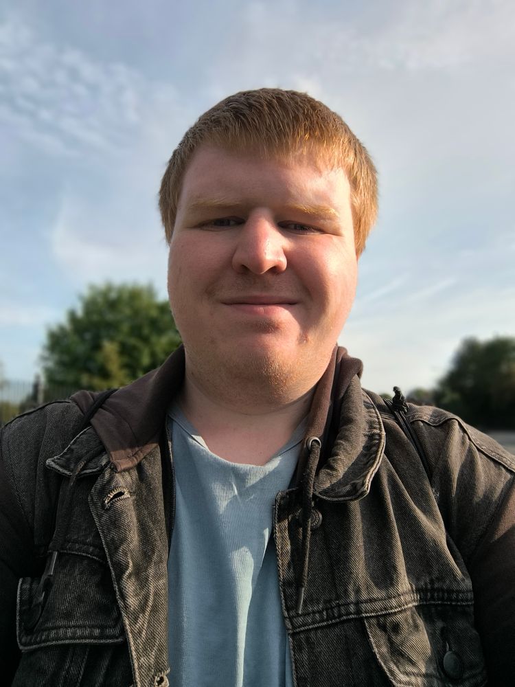 Selfie taken outdoors of a white, masc presenting person with short ginger hair wearing a denim hoodie and a blue shirt underneath