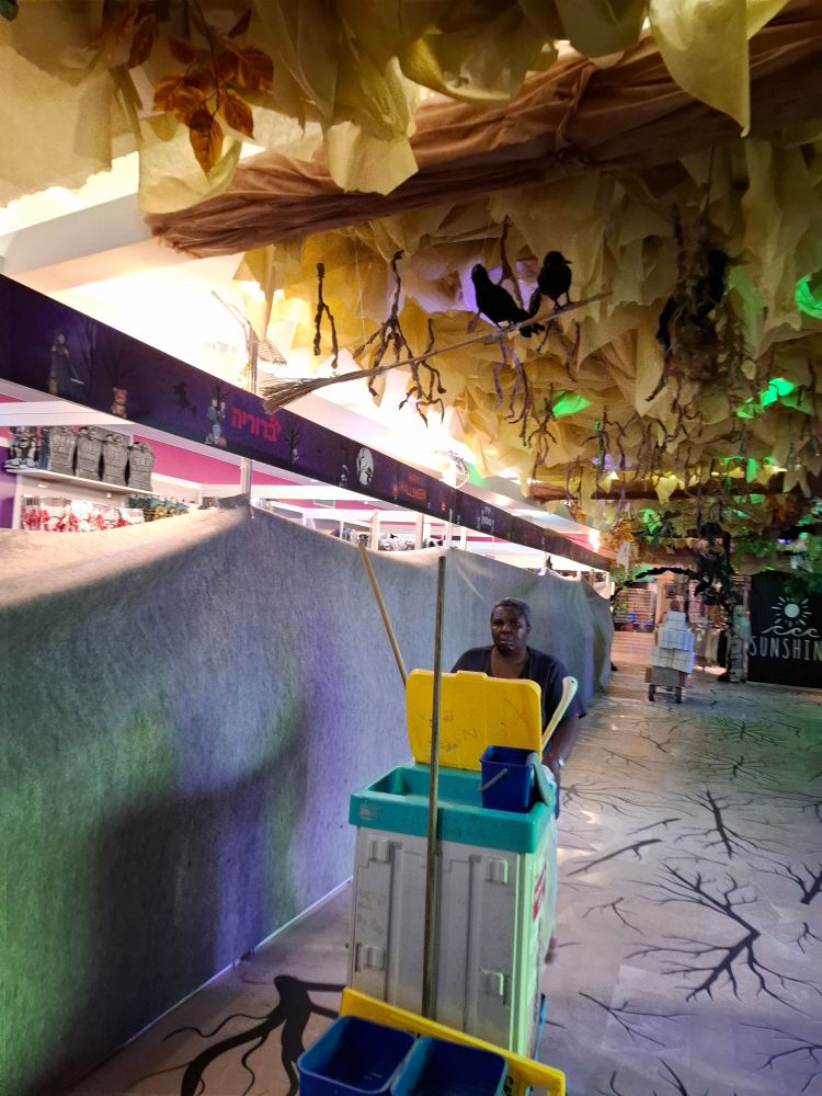 A woman pushing a cleaning cart and a man pushing a two wheeled cart stocked with boxes in a Halloween themed corridor 