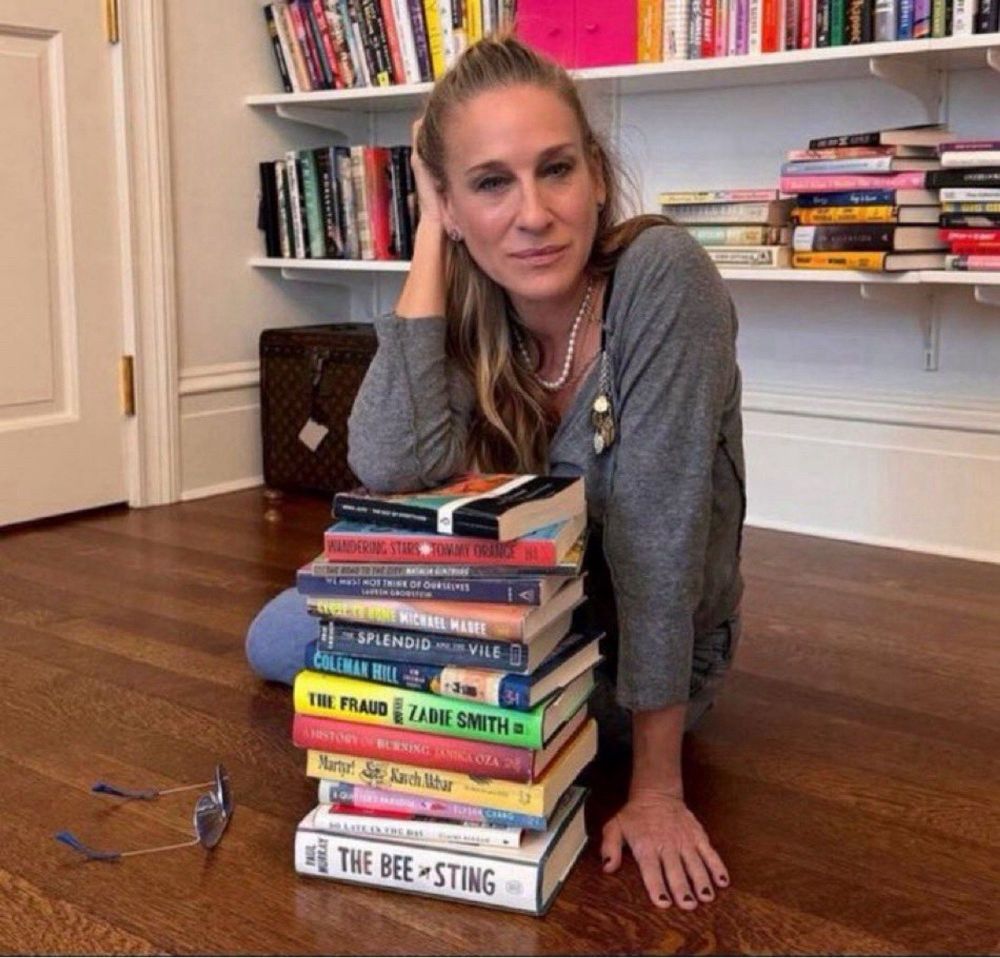 sarah jessica parker sentada no chão se apoiando em uma pilha de 13 livros.

ambiente de casa, chão de madeira óculos no chão. ela está com o braço esquerdo no chão e o outro braço apoiado nos livros com cara de tonta