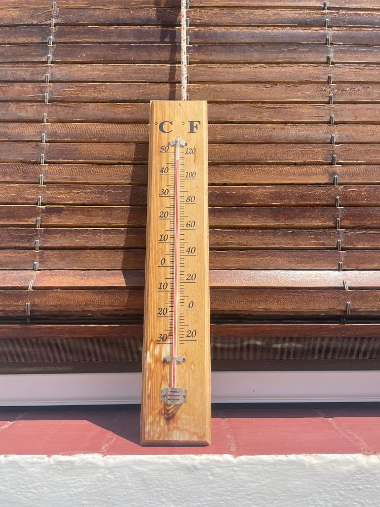 A wooden thermometer is propped up against a wooden blind on a red brick window-sill of a white wall. The gauge reads 47°C