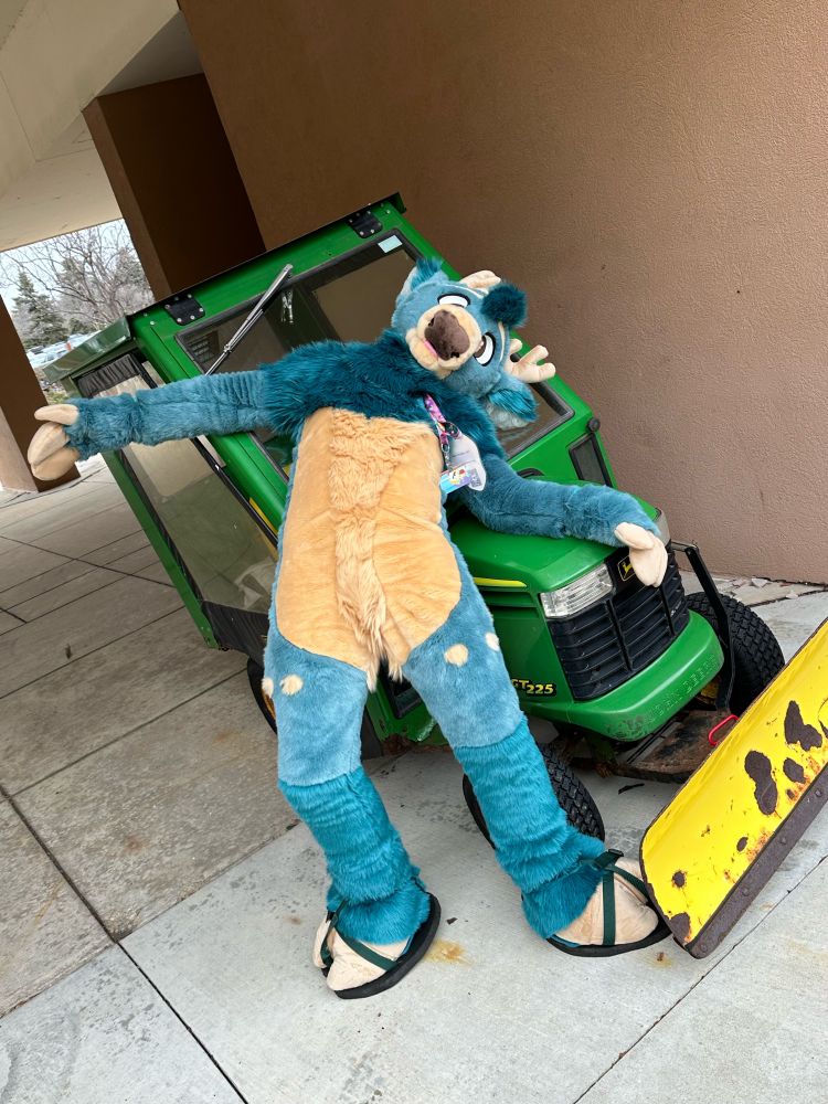 A deer fursuit pretending to be hit by a green tractor. He is laying on his back on the hood.
