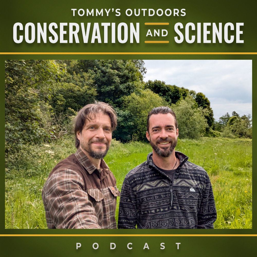 Two men standing in a lush green meadow with dense woodland in the background under an overcast sky. The guest on the left has shoulder-length brown hair and a beard, wearing a brown and cream chequered flannel shirt. The podcast host on the right has short dark hair and a beard, wearing a dark patterned fleece jacket. Both men are smiling at the camera in what appears to be a natural countryside setting with tall grass and mature trees creating a scenic outdoor backdrop.