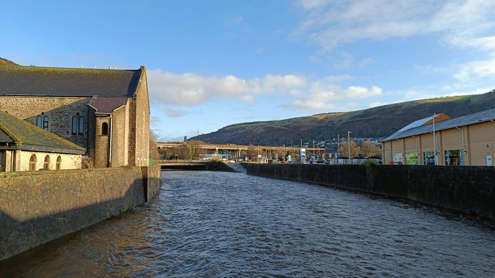 A blue sky over a grey river. A church reaches in from the left. A hill haunts the background in the right. Wales 2025.