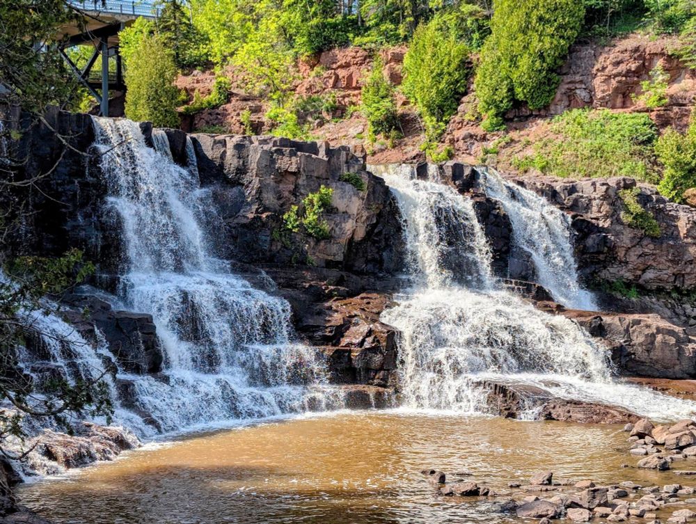 there's a pool of water at the bottom with 3 falls splashing over a rock formation with a bridge in the background