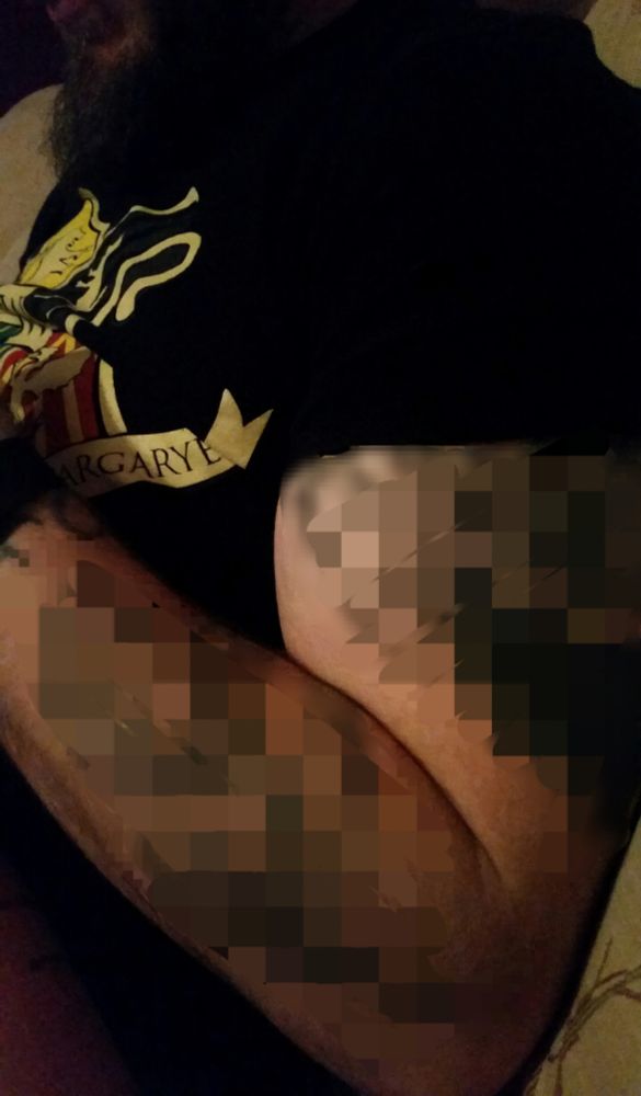 Photo - side on view of my husband as he was sitting next to me. Your pov is from his chin down to his waist. But you mostly only see his arm, shoulders and chest. His left arm is raised bc he has his phone in his hands reading something. There are big and medium sized tattoos on his bicep and forearm that I have blurred. He's wearing a black Game of Thrones t shirt. 