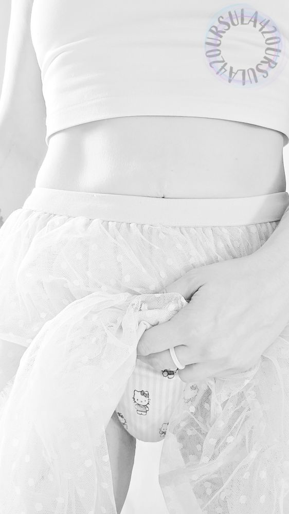 Black and white photo of ursula from mid chest to mid thigh. Her tummy and hips are the centre point of the image. She is using her left hand to lift her skirt a little bit, the sheer fabrics bunched in her hand, because you asked her so nicely to show you her panties. She knows she shouldn't be wearing them