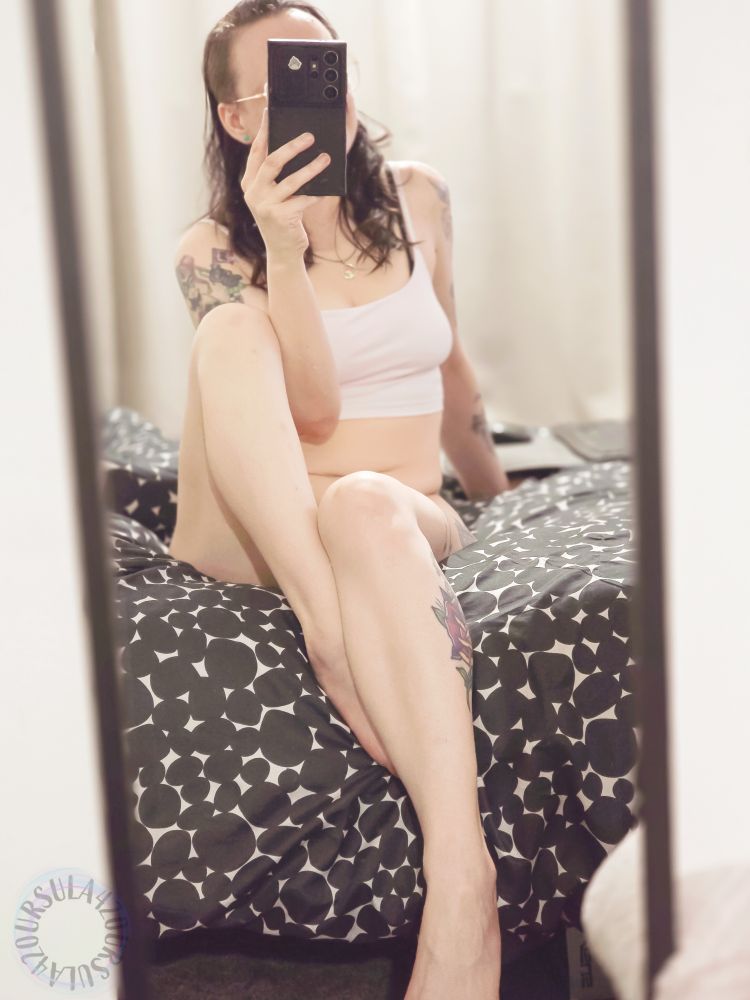 Ursula is sitting on her bed in nothing but a pink crop top. Her hair is messy, and she is leaning back on one hand with her phone in the other to take a selfie. One knee is raised up close to her chest while the other is hanging off the edge of the bed.