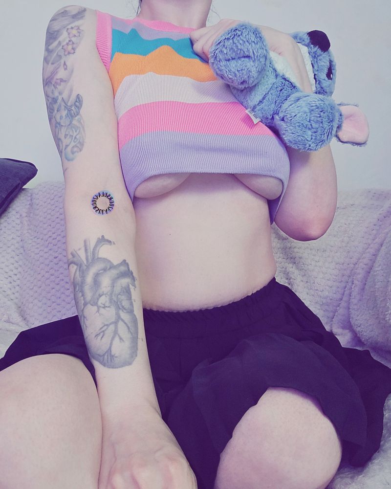 Ursula sitting on her sofa in her black pleated school skirt and a crop top in rainbow stripes. The crop top has been pulled up to give you an underboob display. Her tummy and middrift is on show and bare. 

You can she her knees from where her skirt has pulled up a bit from how she is sitting and she is facing the camera but turned slightly off to her left.

She is holding high on her chest and close to her body a plushie of Stitch from Lilo & Stitch. One of her favourite characters & a thing she has an affinity for. Lilo & Stitch can trigger a bit of flapping with her autism.

A heart tattoo on her right forearm is visible and A large Geisha on her right bicep is also on show.