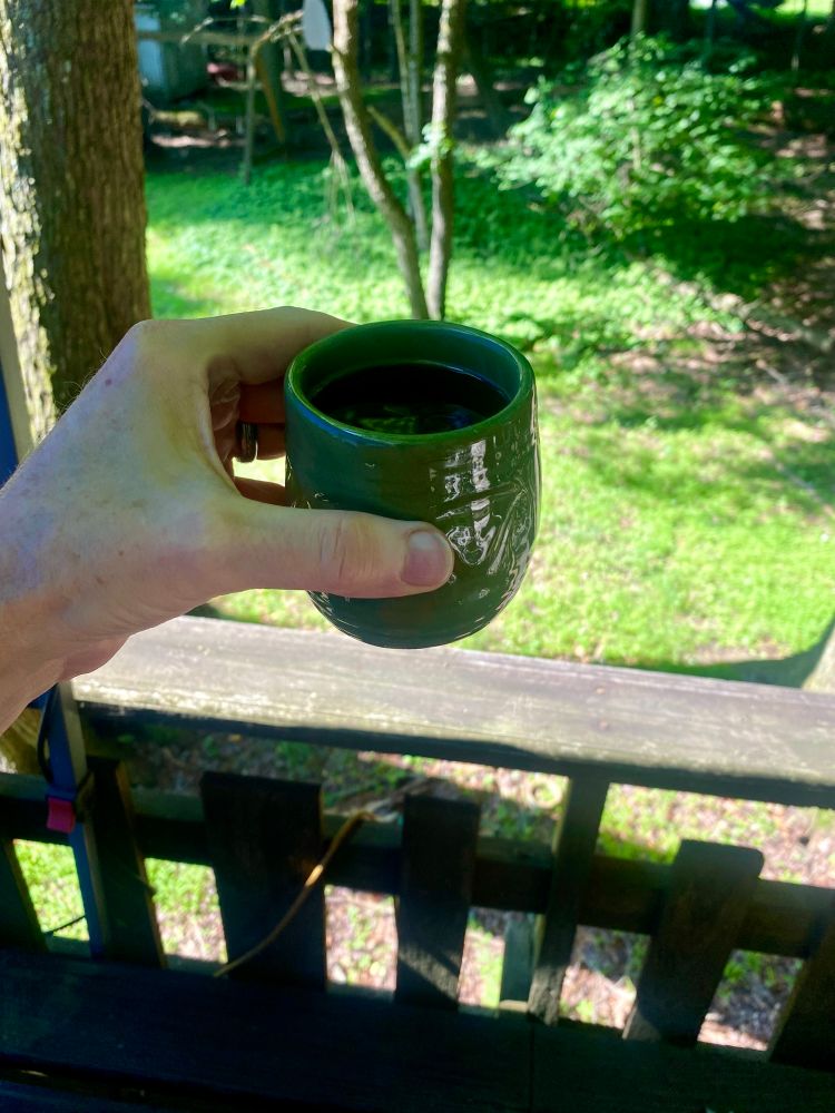 Hand holding up a cup of coffee with deck railing and trees in background. 