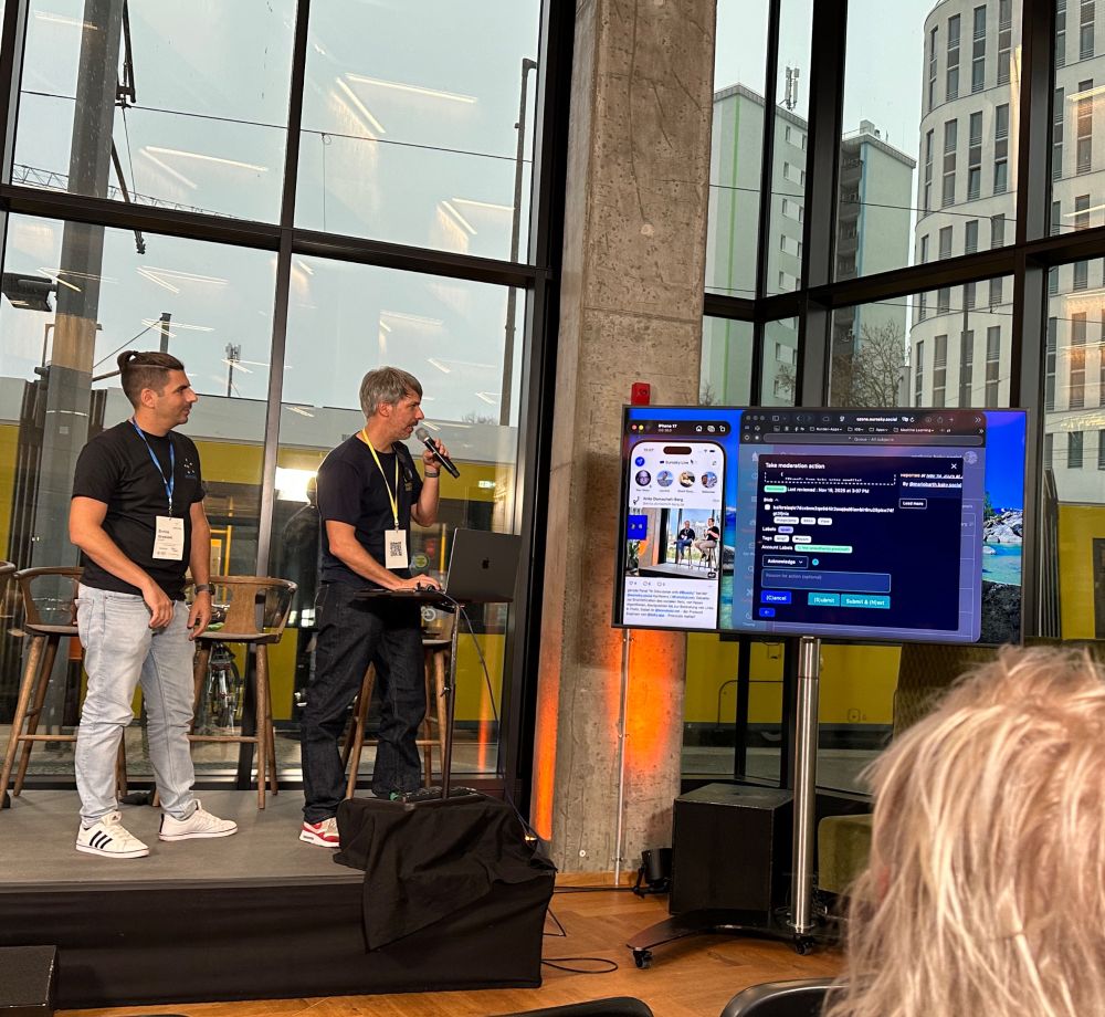 2 Männer stehen auf einer Bühne u gucken auf einen großen Screen. Links auf dem Screen ist mein Skeet eingeblendet. 