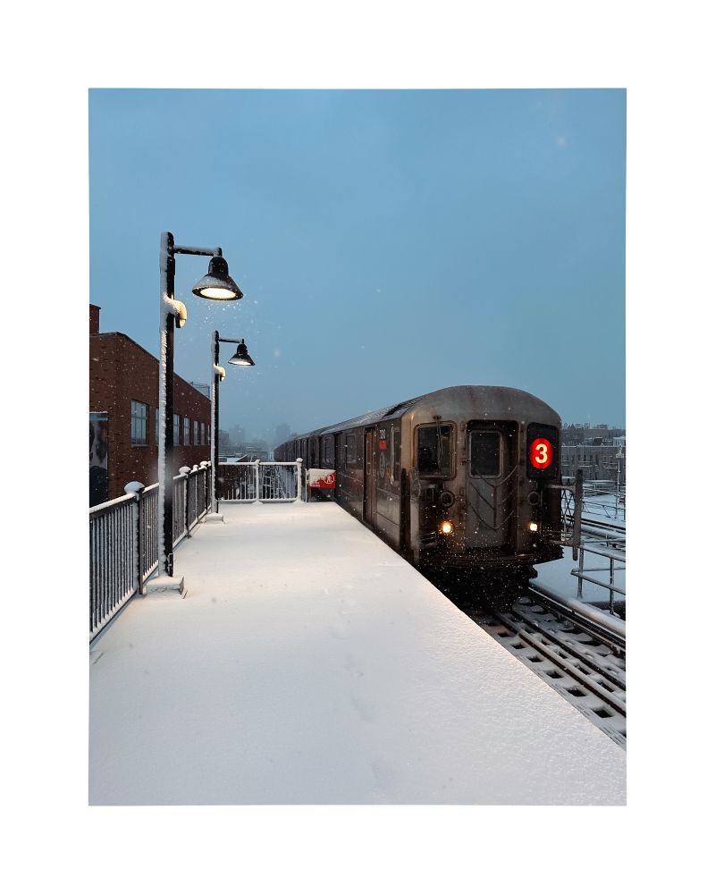 An old silver 3 subway train pulling into an elevated station platform. It’s snowing and the platform is covered with snow. 