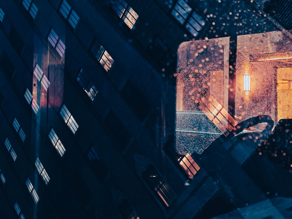 Reflection of a city building on a rain covered windshield. There’s a light from the building across the street shining on the window 