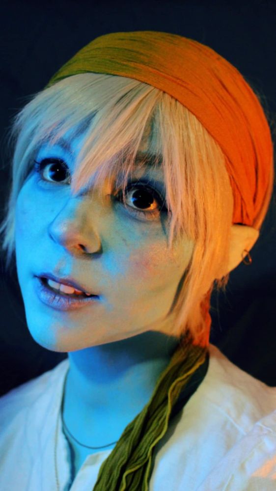 Portrait of a fantasy character with blue skin, large black eyes and white hair. The character is staring off. They are wearing a yellow bandana and a white shirt. 