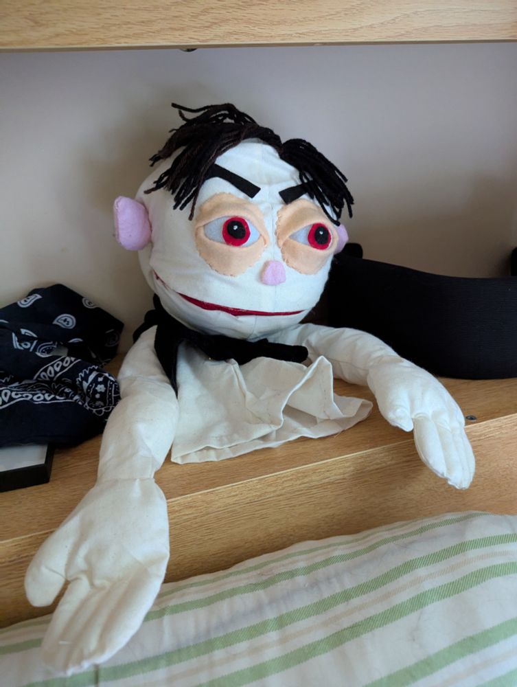 Puppet with fair skin, red eyes, and black yarn hair perched on a shelf. 