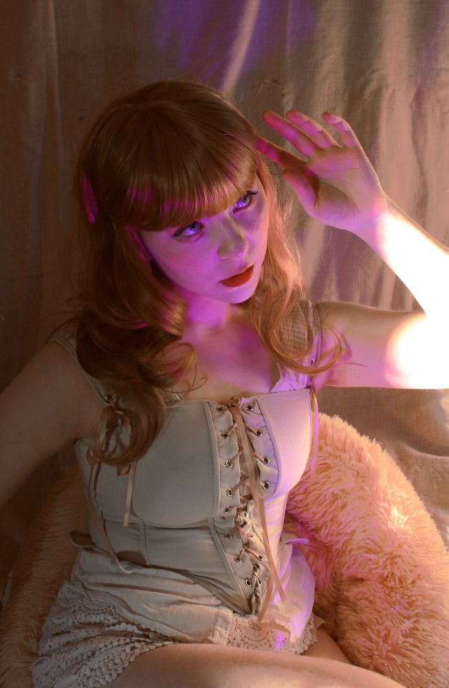 Girl in corset sitting in a fluffy chair. Long golden blonde hair her face in pink light. 