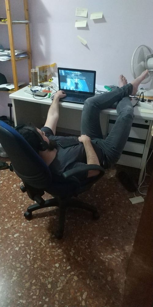 Persona jugant a Hollow Knight en un portatil en la ma esquerra tirat en una cadira en les cames damunt la taula i l'altra mà en el mòvil en la panxa
