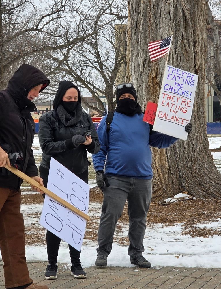 Protesters holding signs. On the left a protester holds a sign reading coup number two on the right a protester is carrying an American flag and a sign reading they're eating the checks they're eating the balances
