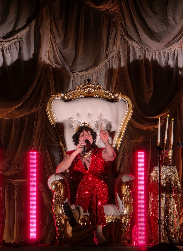 Performer Spencer Sutherland, a white man wearing a red sequin suit, sitting on a throne wearing a golden crown 