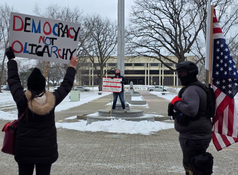 Far left, the back of a protestor holding a sign reading "democracy not oligarchy"; far right, the side of a protestor holding an American flag. They are sandwiching at protestor facing the camera and holding a sign reading "Americans deserve Healthcare"