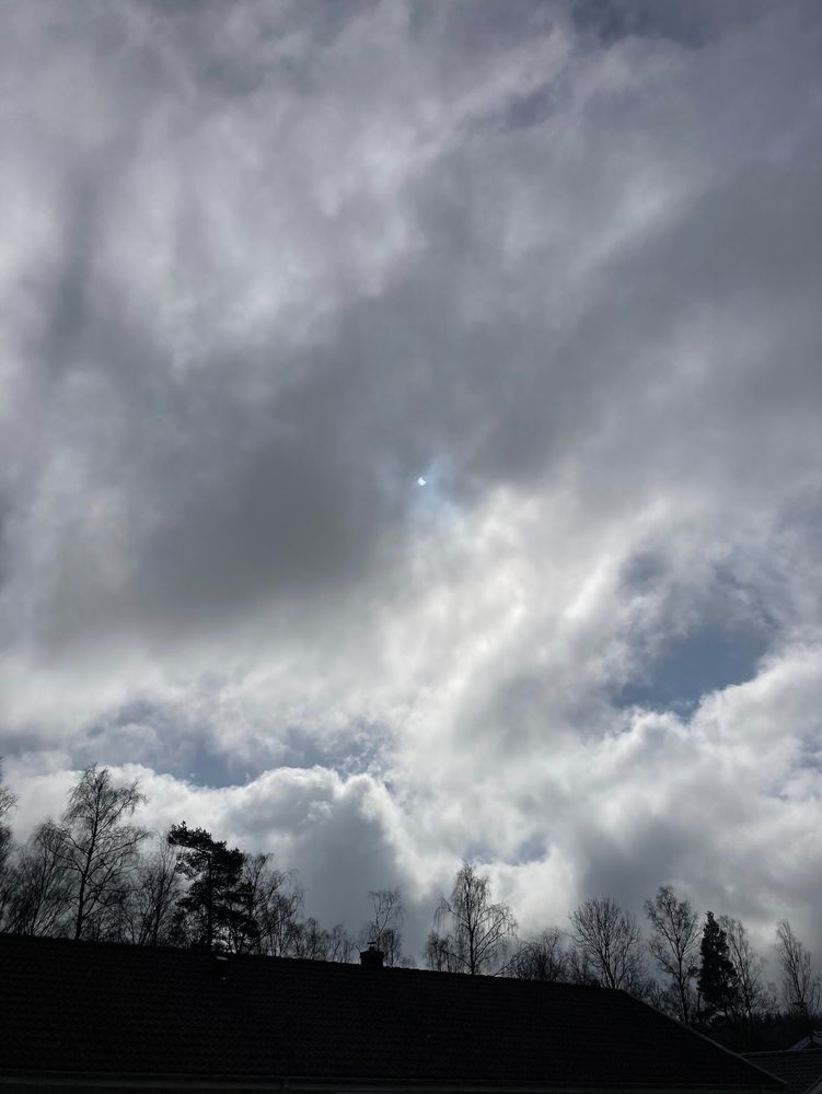 Partial Solar Eclipse, Fotskäl, Sweden. Rooftop, silhouettes of trees, grey-white cloud, small sun in middle. Clouds acting as filter revealing 0.34 eclipse by moon from top right.