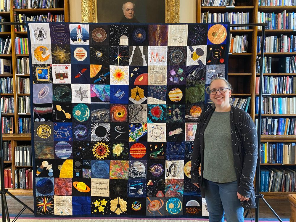  A woman standing by a quilt in a library. The quilt is Astronomy themed patchwork. 