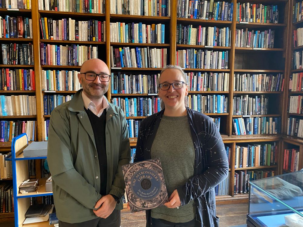 A man and a woman standing in a library holding a book called “The Astronomers Library” 