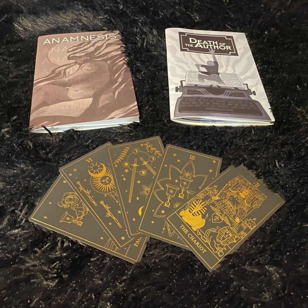 Copies of the solo ttrpgs Death of the Arthur and Anamnesis by Sam Leigh. Below them 5 black and gold tarot cards. From left to right: Page of cups, six of wands, two of swords (reversed), 3 of cups, and The Chariot. 