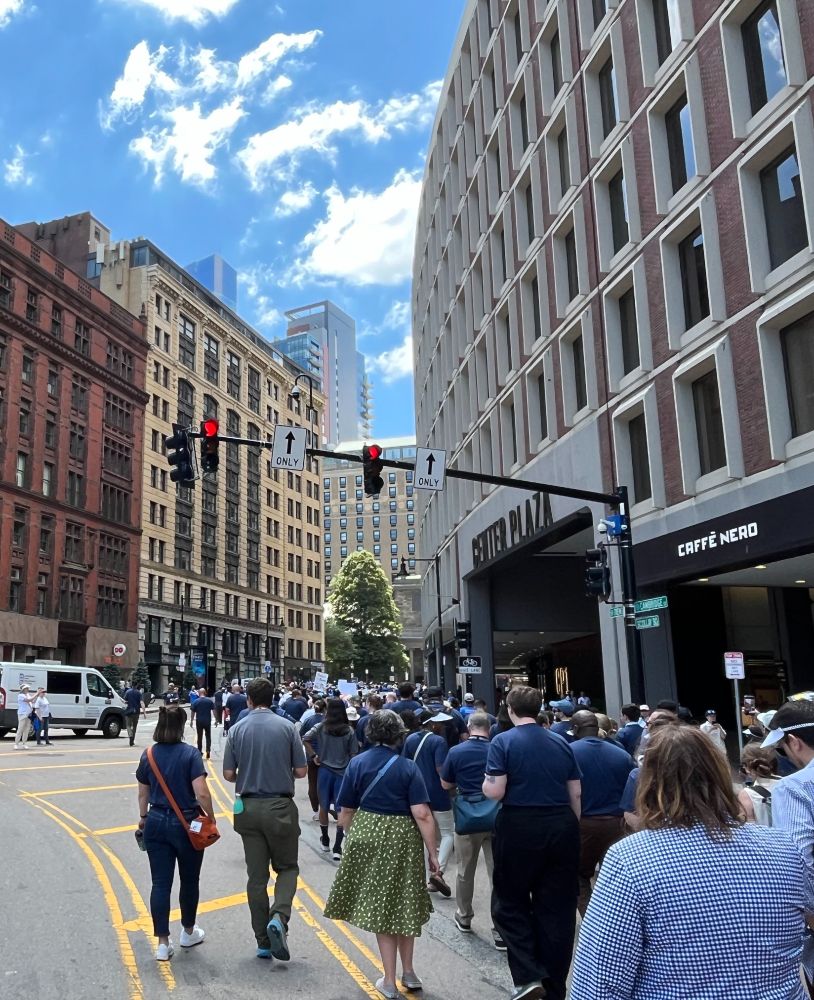 A thick crowd of people march in the middle of Cambridge Street, which transitions into Tremont Street, in Downtown Boston. Most marchers are wearing navy blue shirts. The street is lined with 10–12 story buildings; a Caffè Nero is see at right.