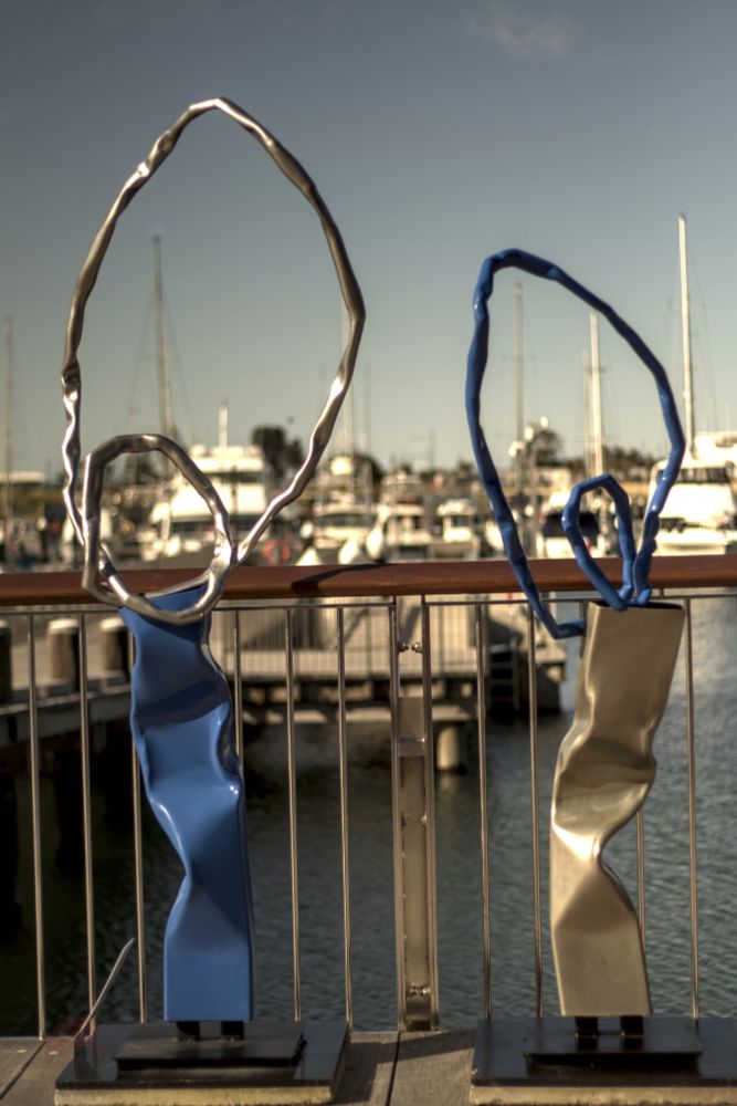 Two sculptures standing side by side.  The first consists of a metal rectangular box that has been slightly bent and deformed. Out of the top of the box, a silver wire loops out to form a large vertical oval, and then a smaller oval within that oval, before reconnecting to the box.  The second is similar to the first except that the colours have been switched, with the supporting metal box being silver, and the wire being blue.
