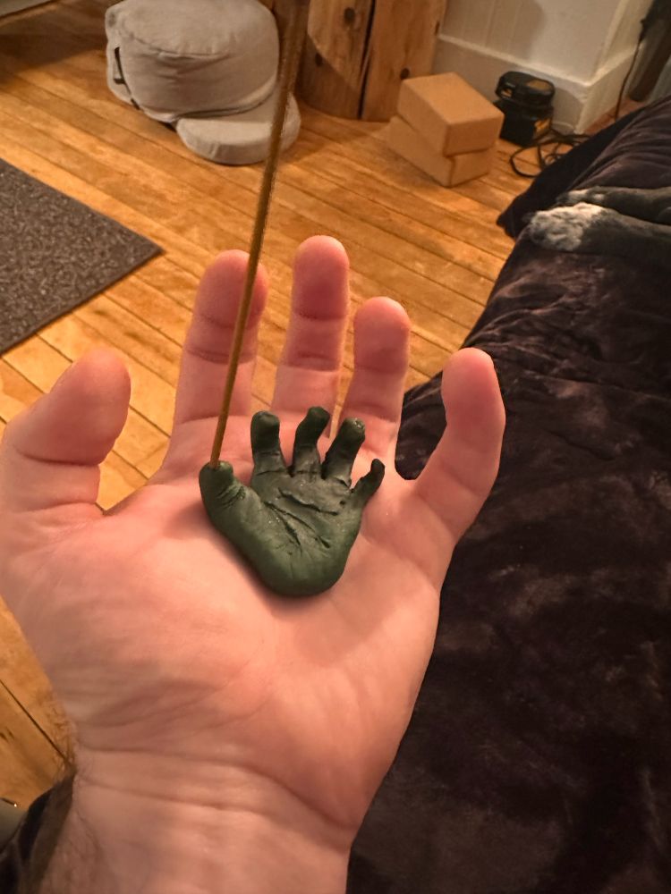 Left hand holding a small green clay model left hand with a stick of incense standing in the thumb of the clay hand.