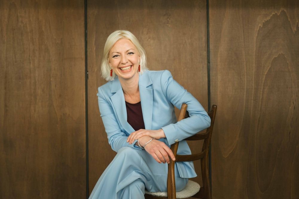 Eine lachende, sitzende, blonde Frau in hellblauem Anzug