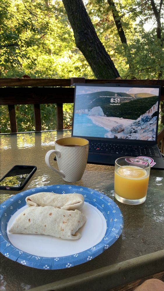 Two breakfast burritos sit on a paper plate that is on a glass patio table ln a deck in the woods. There is a cup of coffee and a glass of orange juice on the table sitting in front of a laptop (locked screen) 