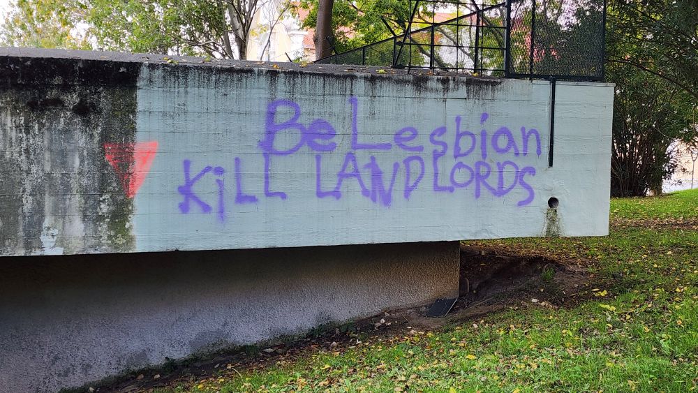 An upside down pink triangle to the left of purple graffiti that says Be Lesbian Kill Landlords