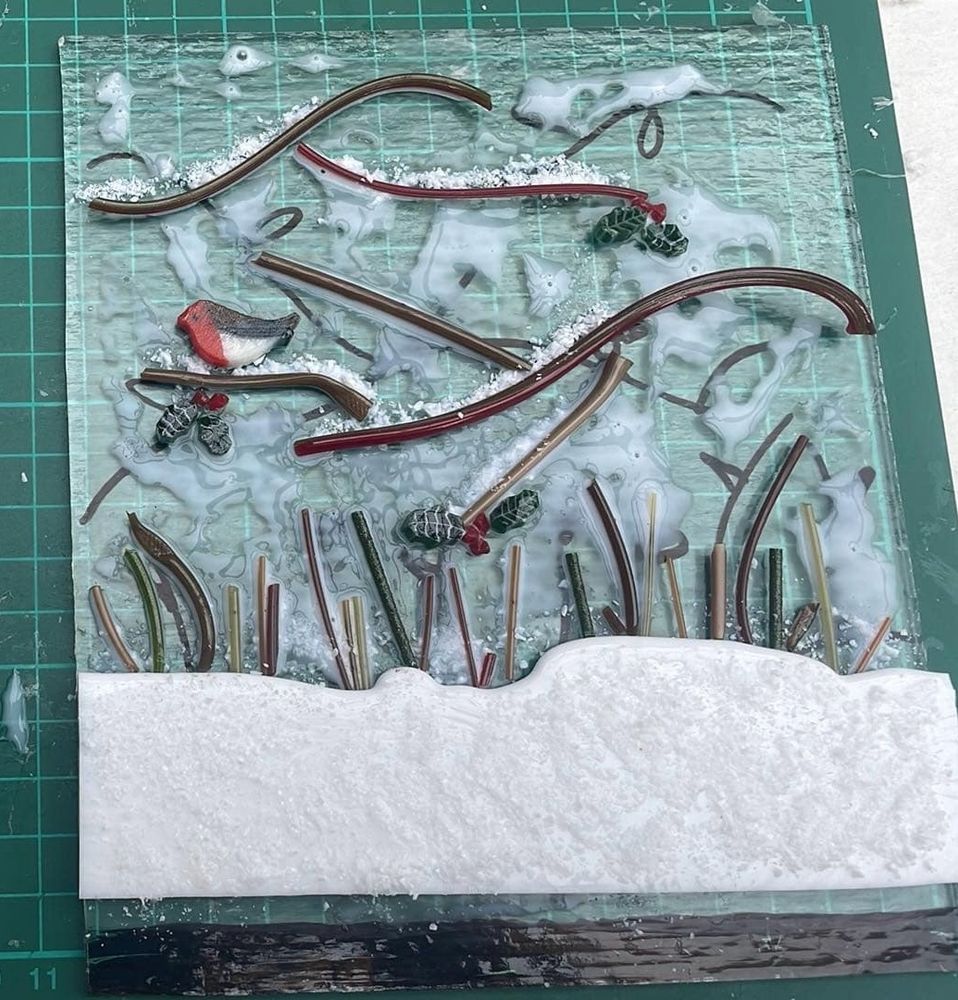 A rectangle of glass, with smaller bits of glass glued to it to make a scene of a robin on a bare wintry branch in the snow