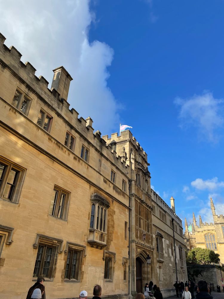 A building in Oxford, on a clear sunny day. It’s grand and long, with battlements, and the trans flag is flying on its flagpole. 