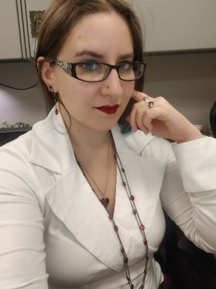 A selfie of Fey sitting in their cubicle. They're wearing a snug, long sleeve white blouse with a wide collar and lapels; a double strand necklace of red gems with matching earrings, and a red heart pendant. Several rings grace their fingers. Narrow rectangular glasses do little to hide their chilly expression, nor detract from their full, blood-red lips