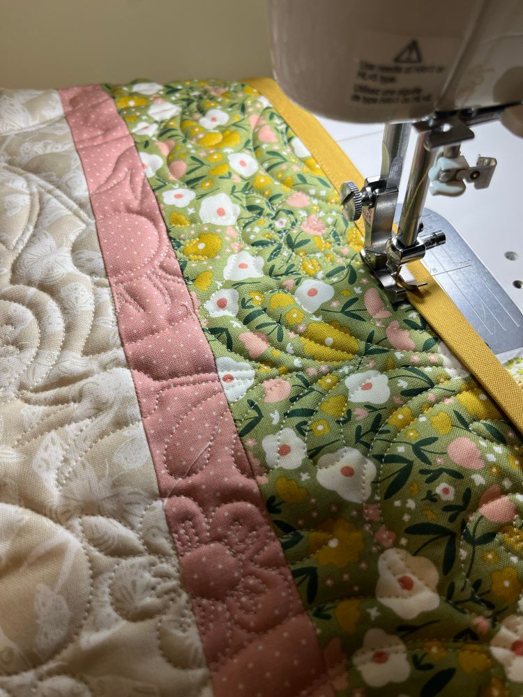 A sewing machine adding mustard yellow binding to a green floral quilt edge.