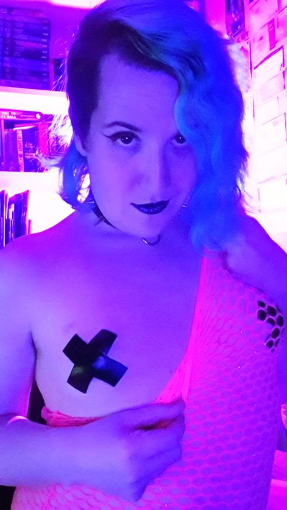 Backlit and in pale purple-blue lighting, I'm pulling down neon-pink fishnets, showing off the black X nipple pasties underneath. My smirk is emphasised by the black lipstick I'm wearing.