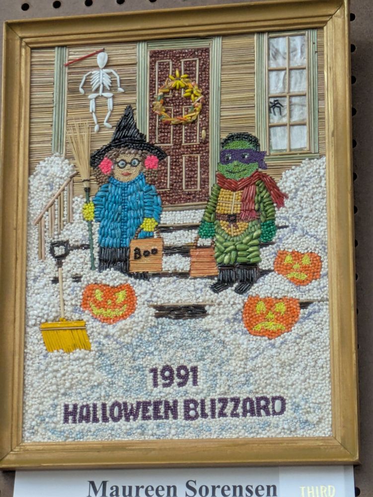 Crop art from the MN state fair 2025. It depicts children in costumes on snowy steps in front of a house. There are Halloween decorations about. There is text at the bottom that says 