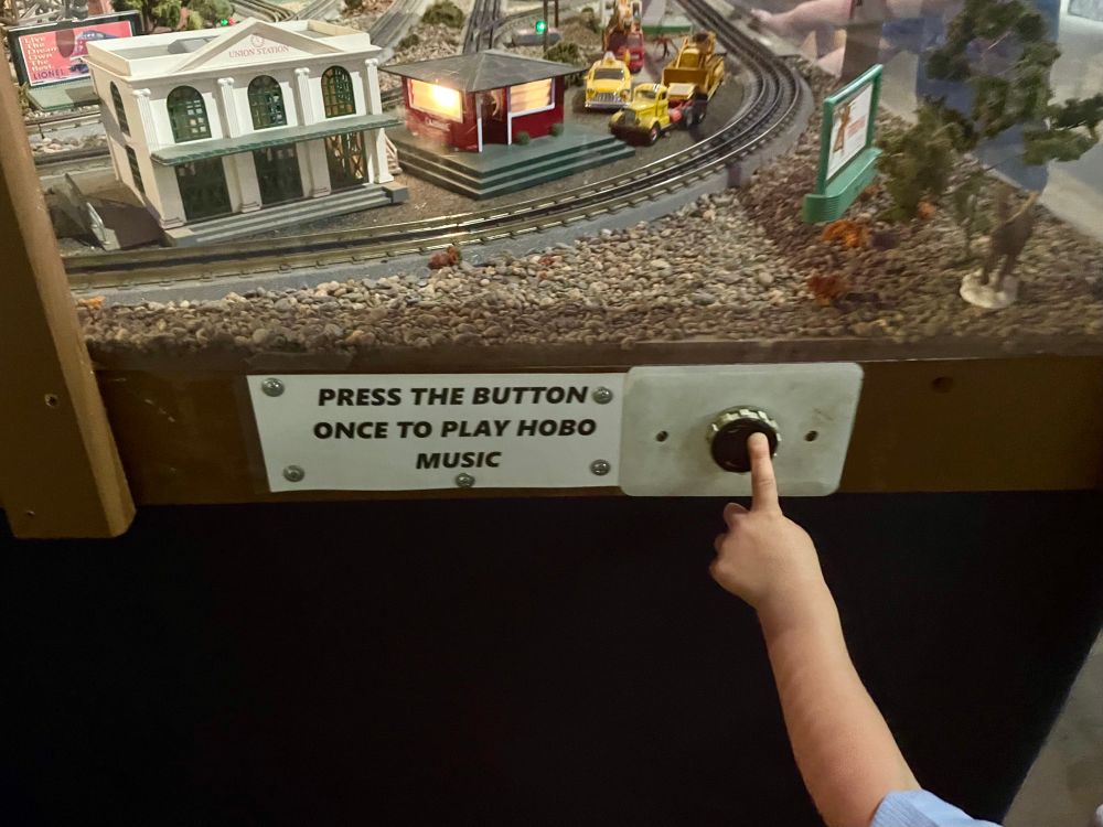 A button on the side of a model train set with the sign “press the button once to play hobo music”, and a child’s hand reaching for the button. 