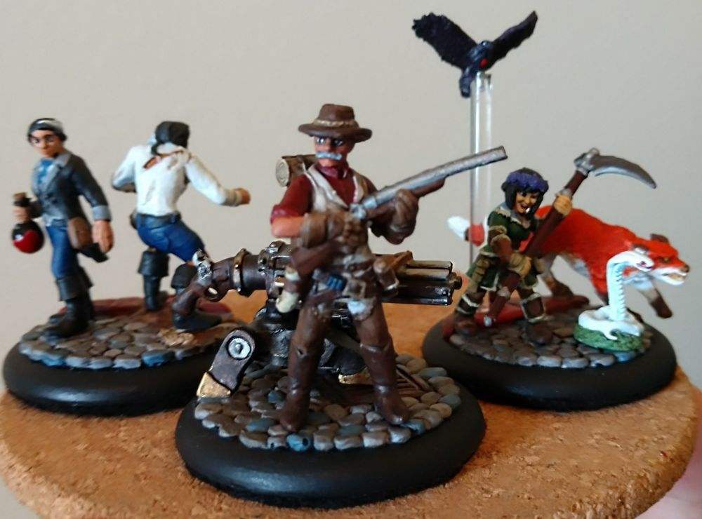 Three painted miniatures of the PCs for a Pathfinder game I ran, displayed on a cork coaster. Left: Odette DeSade, an Alchemist in a blue-black tail coat holding her mutagen in a flask, standing back-to-back with her combat alter-ego. Center: Charles Woodruff, a Gunslinger in a red shirt and cream vest holding his rifle, and standing in front of his custom repeating turret. Right: Mori, a Druid in green hide-armor wielding a scythe, standing next to her animal companions; Fox the dire fox, Snak the albino cobra, and Edgar Allen Crow the crow.