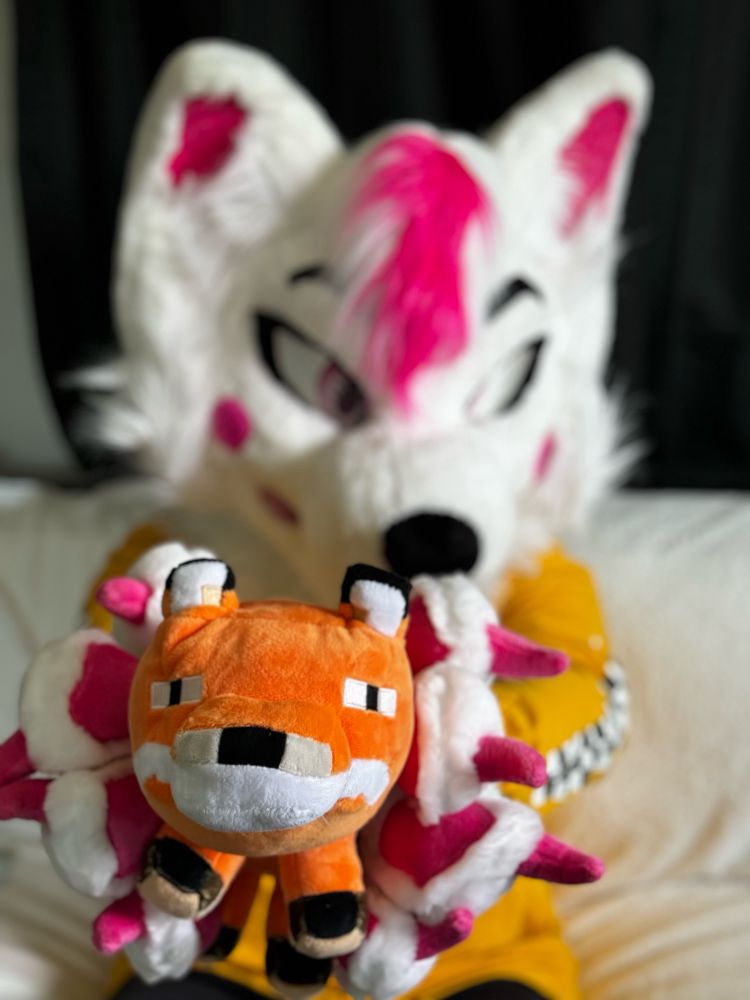 The white pink fox fursuiter extends his arms out to show the Minecraft plushy