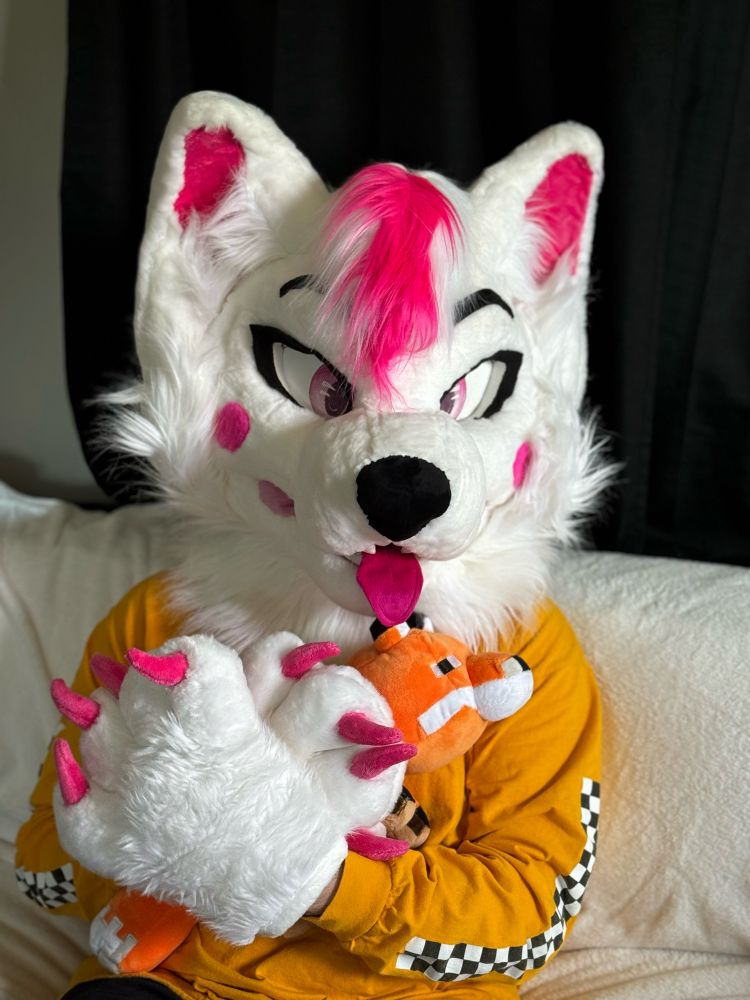 A pink and white fursuit fox sitting on a white couch with a yellow long sleeve hugging a Minecraft fox plushy.
