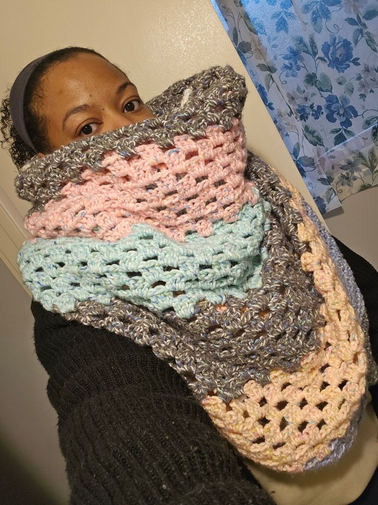 My blanket scarf is thick and long. The part that's showing is  granny stitched and striped with gray, pink, blue, orange, green, and white yarn. It is wrapped on me, ends backward, covering my nose and down to my pelvis. It's wide enough to cover my shoulders. I'm wearing a black cardigan and a tan sweatshirt. My hair is in a ponytail with a grey headband. I'm in a room with a window that has a sheer floral curtain.