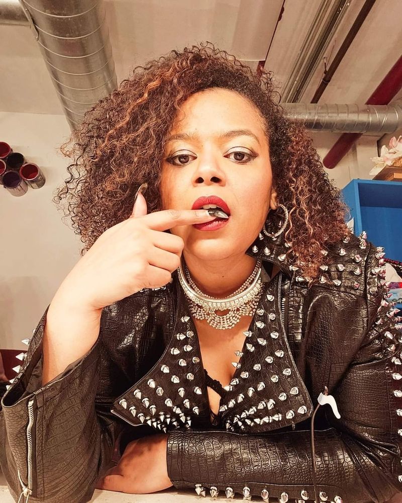 I'm leaning towards the camera, one arm resting on top of a counter, the other arm has the elbow on the top, with my pointer finger with long smoky nails at my lips. I'm wearing red lipstick and my curly brownish-red hair is about shoulder length and fluffy. I'm wearing silver necklaces and chokers, and a black leather jacket with studs. I'm in a room with pipes on the ceilings and walls