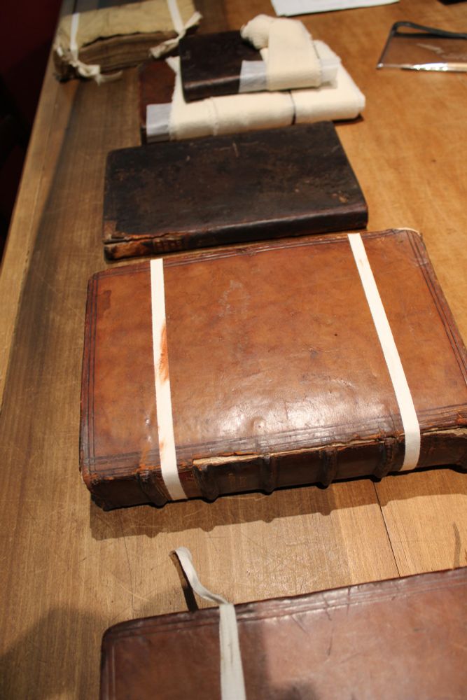 The image shows the length of a table with several books in a row ready to be repaired. At the end of the table are two books where the repair process has begun, and they have been wrapped in bandages. The covers are mostly 17th or 18th century in date.